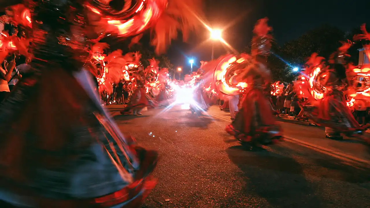 Foliões com fantasias vermelhas rodopiando em desfile noturno numa rua, com o movimento borrado e a luz de um refletor estourando ao fundo