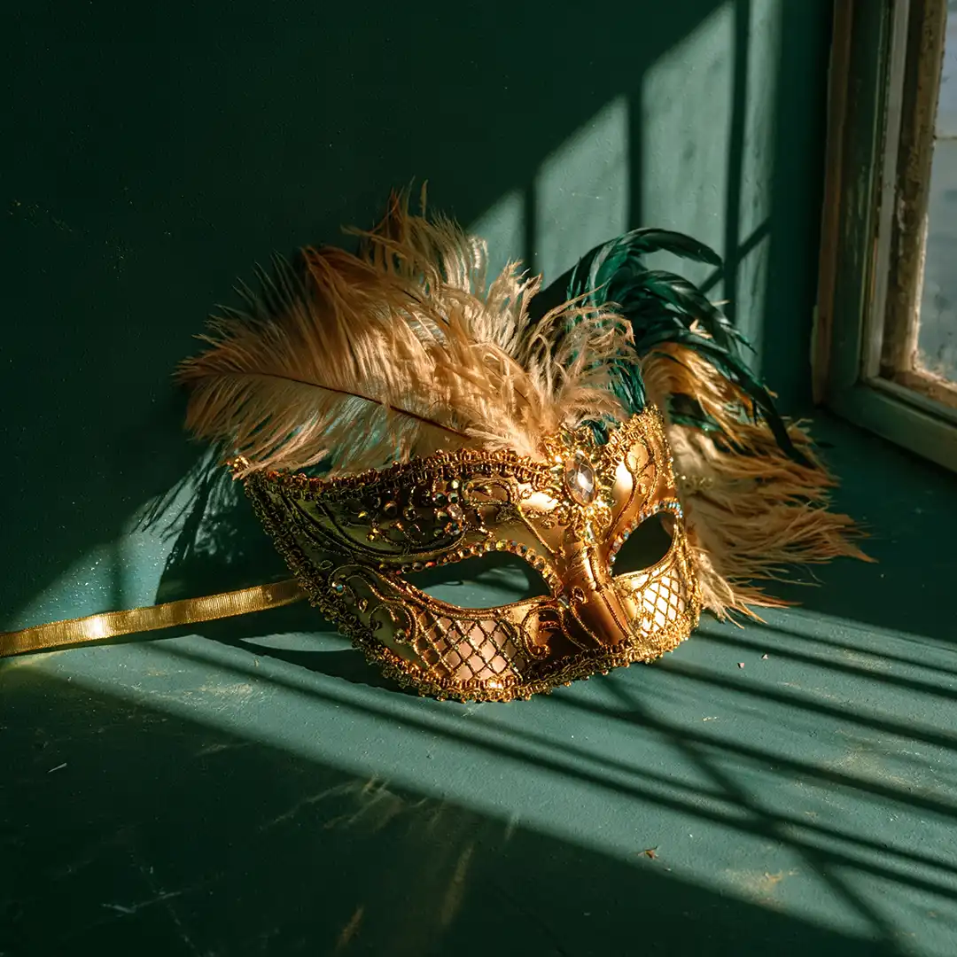 Máscara de carnaval dourada com penas creme e verdes apoiada num peitoril, com sombras de veneziana na parede verde