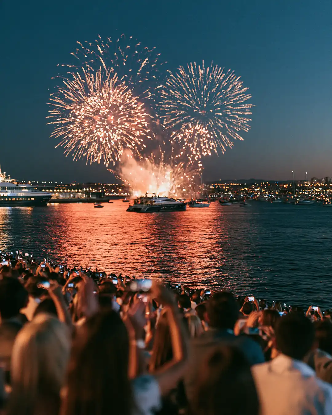 Fogos de artifício explodindo sobre a água enquanto uma multidão de costas assiste e fotografa com celulares